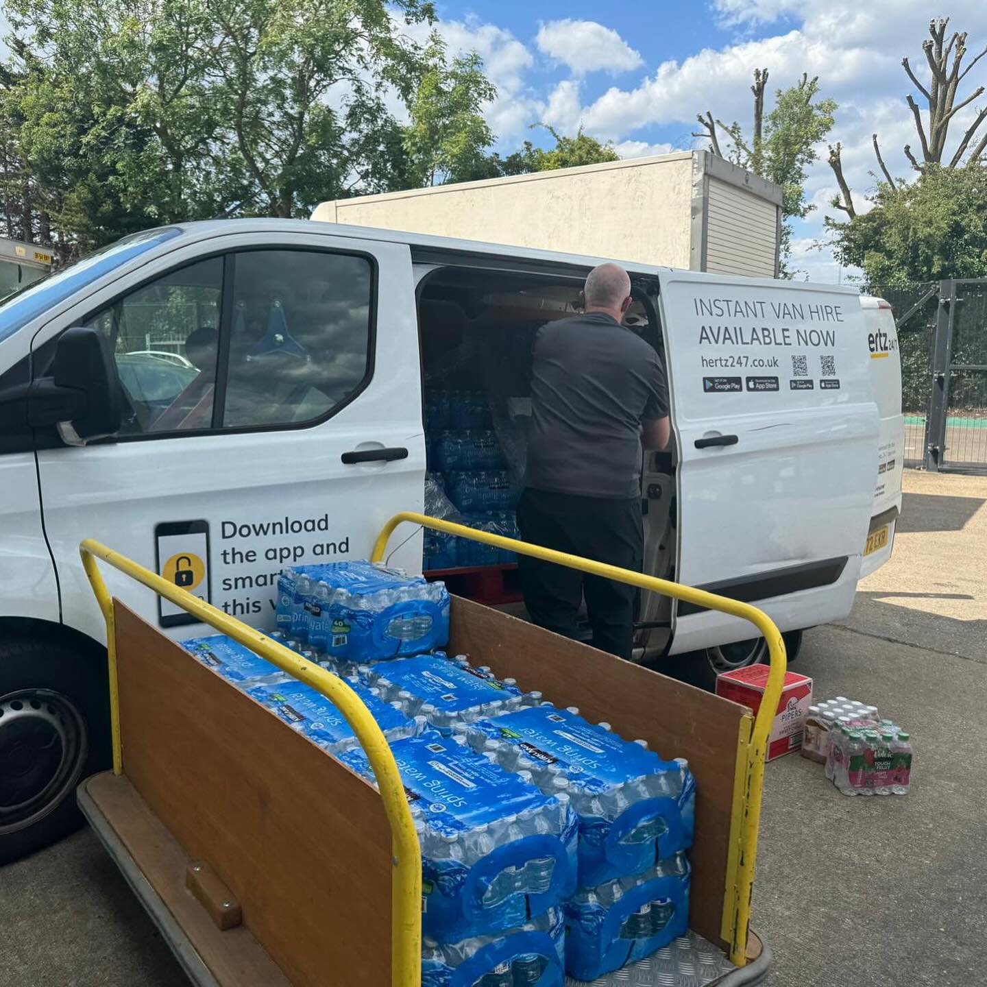 Man unloading packs of Kirkland Signature spring water bottles from a white Hertz 24/7 rental van, with a trolley filled with multiple 40 x 500ml cases.
